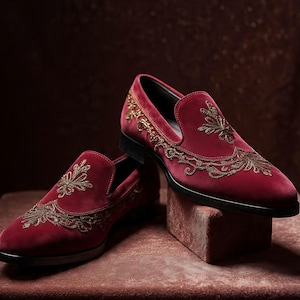 May include: A pair of burgundy velvet loafers with intricate gold embroidery. The slip-on shoes have a classic design with a pointed toe and a black sole. The gold embroidery is in a floral pattern.