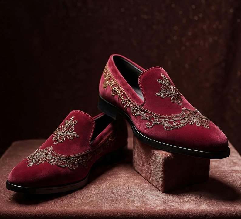 May include: A pair of burgundy velvet loafers with intricate gold embroidery. The slip-on shoes have a classic design with a pointed toe and a black sole. The gold embroidery is in a floral pattern.