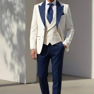 May include: A three-piece suit in ivory with navy blue accents. The suit includes a jacket with navy lapels, a vest, and navy trousers. The outfit is completed with a navy tie and brown leather loafers. The man is smiling.