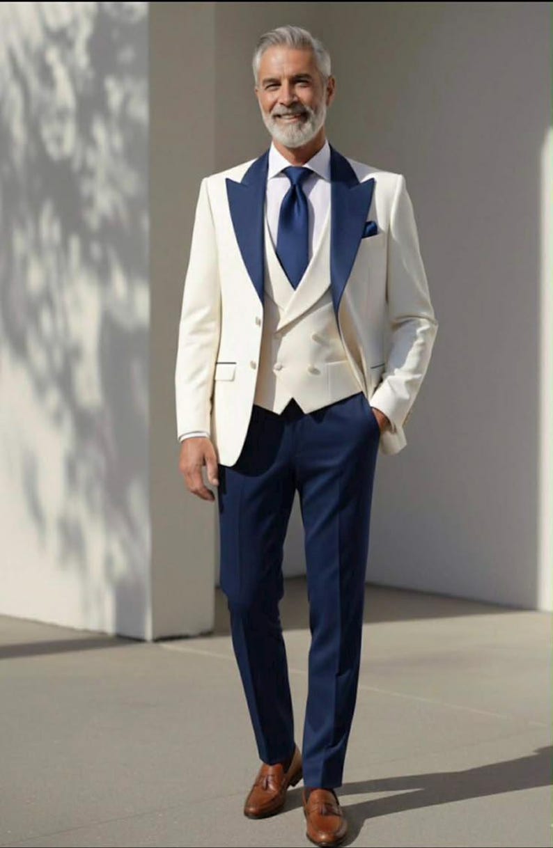 May include: A three-piece suit in ivory with navy blue accents. The suit includes a jacket with navy lapels, a vest, and navy trousers. The outfit is completed with a navy tie and brown leather loafers. The man is smiling.