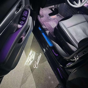 Puede incluir: Vista interior de un coche Cadillac con elementos iluminados. El umbral de la puerta muestra el logotipo y el nombre de Cadillac en luz blanca. La iluminación de acento púrpura y azul es visible en todo el interior, incluido el panel de la puerta y las alfombrillas.