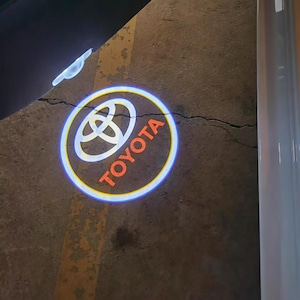 Puede incluir: Un logotipo de Toyota proyectado ilumina una superficie de hormigón. El logotipo es un emblema blanco dentro de un círculo, con la palabra "TOYOTA" en naranja. Un contorno azul y amarillo rodea el logotipo, creando una pantalla vibrante.