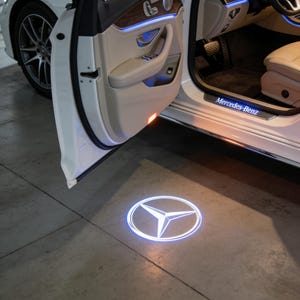Könnte beinhalten: Eine weiße Mercedes-Benz Autotür ist geöffnet und gibt einen cremefarbenen Innenraum mit blauer Akzentbeleuchtung frei. Ein Logo wird auf den Boden projiziert, mit dem Namen Mercedes-Benz auf der Einstiegsleiste.
