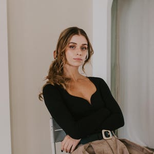 May include: A black long-sleeve top with a deep V-neckline. The top is paired with a brown skirt and a black belt with a gold buckle. The person is seated on a chair in a room with a mirror.