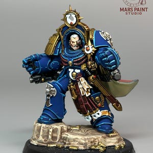 May include: A detailed miniature figure of a space marine in blue and gold armor, standing on a rocky base. The figure has a stern expression and is holding a weapon. The figure is a Warhammer 40,000 miniature. The Mars Paint Studio logo is in the background.