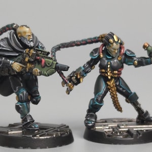 May include: Two miniature figures of futuristic soldiers. One figure is dressed in black with a cape and holding a green and gold weapon. The other figure is in black and teal armor, holding a green weapon. Both figures are on black bases.