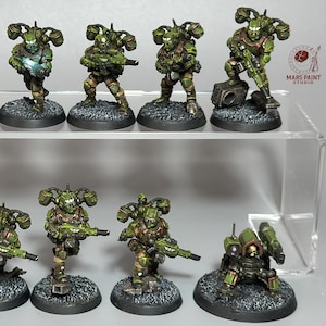 May include: A collection of miniature figurines, primarily in shades of green and brown, depicting futuristic soldiers. Each figure is mounted on a small, textured base. The figurines are arranged on a clear display shelf, with the Mars Paint Studio logo visible.
