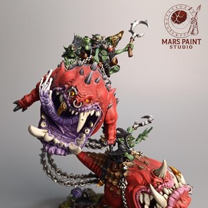 May include: A detailed miniature figure of a fantasy creature with a red body, purple mouth, and sharp teeth. Several green goblin-like figures ride on top. The figure is mounted on a base with miniature plants and mushrooms.