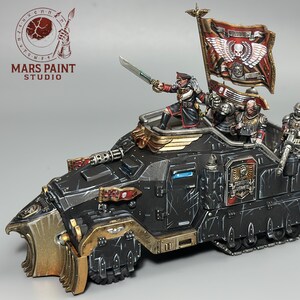 May include: A painted miniature armoured vehicle featuring a black hull with gold trim, a front plough, and a large flag. An officer figure stands on top holding a sword, accompanied by two crew members operating a mounted heavy weapon on the rear deck.