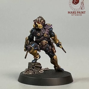 Puede incluir: Figura en miniatura de un guerrero futurista en cuclillas. La figura está pintada en tonos azul oscuro, dorado y bronce, con una máscara de gas y dos pequeños cuchillos. La base es negra y la figura está sobre una plataforma detallada.