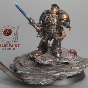 May include: A detailed miniature figure of a warrior in dark armor, holding a blue-bladed sword. The figure stands on a rocky base with red and gray tones. The figure has gold accents and a skull emblem. The text "MARS PAINT STUDIO" is visible.