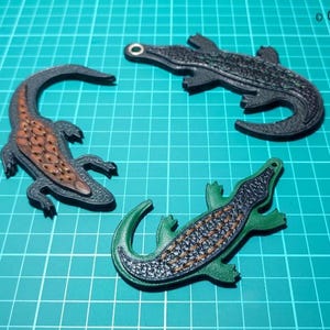 May include: Three alligator-shaped leather ornaments in black, green, and brown, arranged on a teal grid surface. The alligators have textured scales and are designed with a hole for hanging. The image includes the text "Crafted Lines Ideas".