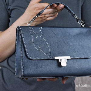 Könnte beinhalten: Dunkelblaue Handtasche mit Klappenverschluss und silbernem Verschluss. Die Tasche hat ein weißes, aufgesticktes Eulenmuster auf der Vorderseite und einen Kettenhenkel. Die Tasche wird von einer Person in einem grauen Oberteil gehalten.