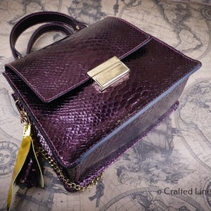 Peut inclure: Un sac à main prune avec un motif texturé en peau de serpent. Le sac est doté d'un fermoir doré, d'une bandoulière en chaîne et d'un pompon. Il possède une poignée supérieure et une fermeture à rabat. Le fond est une carte vintage.