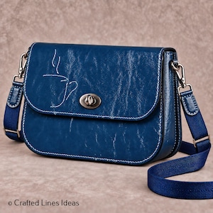 Puede incluir: Un bolso bandolera de color azul intenso con acabado texturizado y el contorno de una taza de café bordado en blanco en la solapa frontal. El bolso cuenta con un cierre giratorio de metal plateado, costuras blancas en contraste y una correa ajustable.