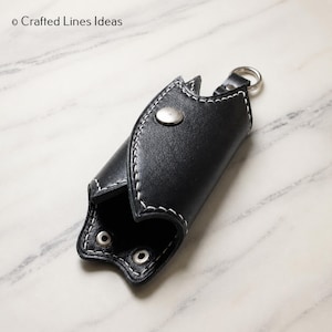 Può includere: Un portachiavi in pelle nera con cuciture bianche a contrasto, una chiusura con bottone a pressione in metallo argentato e un anello metallico circolare. Il testo Crafted Lines Ideas è visibile nell'angolo in alto a sinistra.