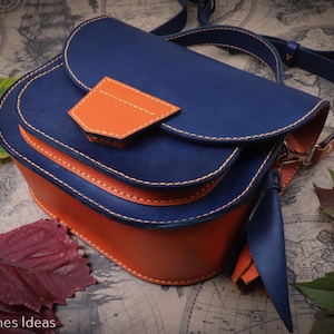 May include: A handcrafted leather crossbody bag in navy blue and orange. The bag features a flap closure with an orange accent, white stitching, and an adjustable strap. The bag is set against a map background with leaves.