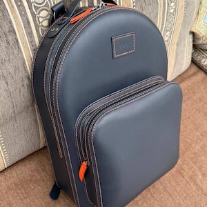 Puede incluir: Una mochila azul marino con costuras y detalles en naranja. La mochila tiene una parte superior redondeada, un bolsillo frontal y un asa superior. Las cremalleras y las costuras son de color naranja, lo que contrasta con el cuero azul oscuro.