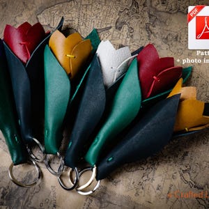 以下が含まれることがあります： 紫、赤、黄色、白などの革製のチューリップのキーホルダーのコレクション。緑の葉が付いています。各キーホルダーにはシルバーのリングが付いています。画像には「Pattern + photo instructions」というテキストのPDFアイコンが含まれています。