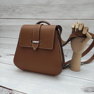 May include: A brown leather backpack purse with a flap closure and a silver-tone clasp. The bag has a top handle and adjustable shoulder straps. White stitching accents the edges of the bag. The background is a light wood surface.