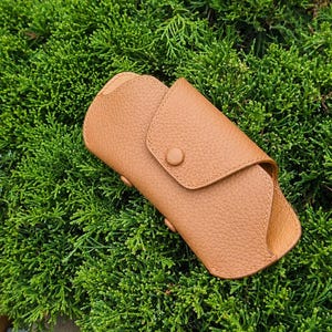 May include: A tan leather glasses case with a textured surface, featuring a rounded flap closure secured by a single matching button. The case has stitched edges and a curved silhouette designed to hold eyewear, with the text Crafted Lines Ideas.