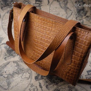 May include: A brown leather handbag with a crocodile skin texture. The bag has two handles and a shoulder strap. The bag is sitting on a map.