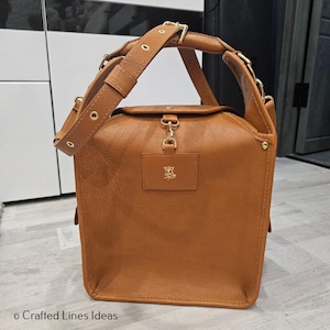 May include: A tan leather handbag with a top handle and a detachable shoulder strap. The bag has gold-coloured hardware, including a clasp closure and rivets. A small rectangular patch with a logo is on the front. The bag is on a light-coloured wooden floor.