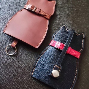 May include: Two cat-shaped leather keychains. One is brown with a matching belt and silver buckle, the other is black with a red belt and silver button. Both have stitched edges and a key ring.