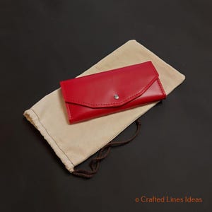 May include: A red leather wallet with a flap closure and a silver snap button, resting on a beige drawstring pouch. The wallet is rectangular and appears to be new. The background is a solid black.