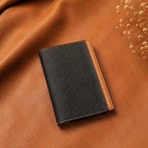 May include: A rectangular bi-fold wallet featuring a textured black leather exterior on the main panel and a contrasting tan leather side panel. The wallet is stitched with dark thread and displays the text Crafted Lines Ideas in the corner.