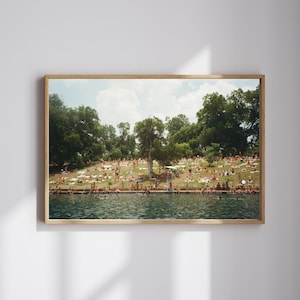 May include: A framed print depicting a sunny day at a lake. People are sunbathing on a grassy hill and swimming in the water. The image has a vintage, film-like aesthetic with a wooden frame.