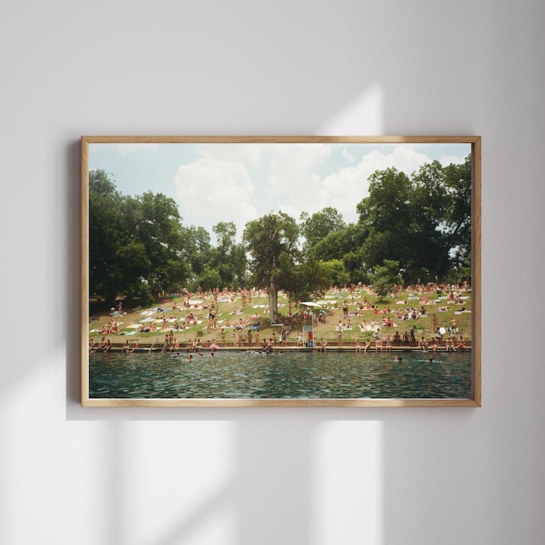 Puede incluir: Una impresi&oacute;n enmarcada de un d&iacute;a soleado en un lago. La gente toma el sol en una colina cubierta de hierba y nada en el agua. La imagen tiene una est&eacute;tica vintage, tipo pel&iacute;cula, con un marco de madera.