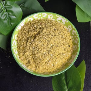 May include: A round ceramic bowl with a green and white floral pattern filled with a coarse, yellow-toned spice blend. The mixture contains fine granules, small dark specks, and tiny white particles, creating a textured, dry appearance.