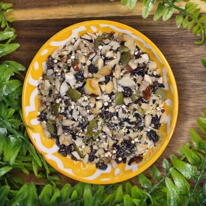 May include: A round yellow bowl with a white decorative pattern filled with a mixture of dried ingredients including dark raisins, green pumpkin seeds, sunflower seeds, chopped nuts, and small grains, viewed from directly above.