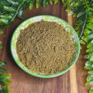 May include: A small, round bowl with a green rim and white floral pattern, filled with a pile of finely ground, light brown powder. The bowl sits on a wooden surface, with green foliage in the background.