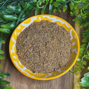 May include: A yellow ceramic bowl with a white patterned rim, filled with a brown granular substance. The bowl sits on a wooden surface, surrounded by green leafy plants. The contents of the bowl appear to be spices or a similar food product.