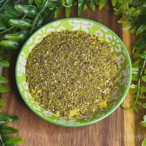 May include: A green and white patterned bowl filled with a granular, light green mixture. The bowl is on a wooden surface, surrounded by green foliage. The mixture appears to be a spice blend or seasoning.