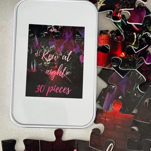 May include: A white rectangular tin containing a jigsaw puzzle with the text "Kew at night" and "30 pieces" in pink. Scattered puzzle pieces show a night scene with purple and red tones. The puzzle is a fun activity.