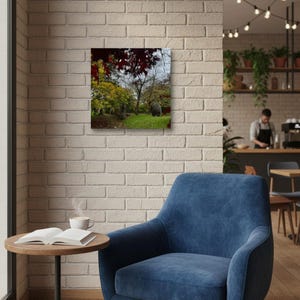 Puede incluir: Fotografía enmarcada de un paisaje de parque con árboles y hierba verde, montada en una pared de ladrillos blancos. Un sillón azul y una pequeña mesa de madera con un libro y una taza de café están en primer plano.