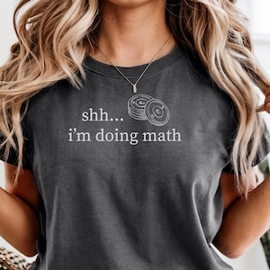 May include: Dark gray t-shirt with the text "shh... i'm doing math" in white. The shirt features a graphic of stacked circles. The model is wearing blue denim shorts and a gold necklace.