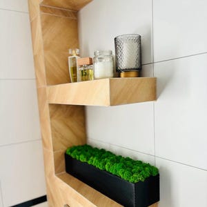 Może przedstawiać: Nowoczesna półka łazienkowa z czarną prostokątną donicą wypełnioną jasnozielonym mchem. Powyżej półka z butelką perfum, małym słoiczkiem i świecą w teksturowanym szklanym świeczniku. Półki wykonane są z jasnego drewna.