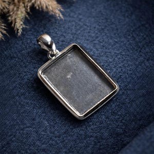 Puede incluir: Un colgante rectangular plateado con un interior gris oscuro. El colgante tiene un lazo en la parte superior para una cadena. El colgante está sobre un fondo de tela azul oscuro.