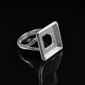 May include: A silver-toned metal ring featuring a square-shaped bezel setting with a hollow center. The band of the ring splits into two distinct sections as it connects to the square frame, creating a polished and reflective surface throughout.