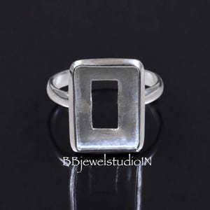 Könnte beinhalten: Ein silberner Ring mit einer rechteckigen Fassung mit hohler Mitte. Das Metall hat eine glatte, polierte Oberfläche. Der Text BBjewelstudioIN ist am unteren Bildrand zu sehen, zentriert unter der Ringschiene und der Fassung.