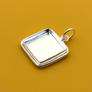 Könnte beinhalten: Ein silberfarbener Anhänger mit quadratischer Lünettenfassung und einer Schlaufe für eine Kette. Der Anhänger hat eine glänzende, reflektierende Oberfläche und ist vor einem gelben Hintergrund platziert. Die Lünette ist für einen quadratischen Stein oder Einsatz konzipiert.