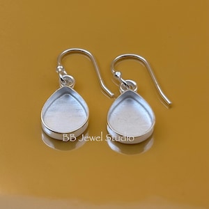 Peut inclure: Une paire de supports de boucles d'oreilles argentés en forme de goutte d'eau. Les boucles d'oreilles ont un sertissage à lunette et une fermeture à crochet. Le texte "BB Jewel Studio" est imprimé au bas de l'image.