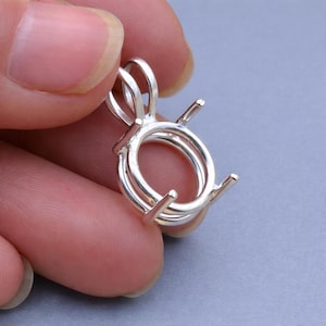 Può includere: Un castone per pendente color argento con castone rotondo e quattro griffe. Il castone ha due anelli per una catena ed è tenuto dalle dita di una persona. Il castone del pendente è progettato per contenere una pietra preziosa.