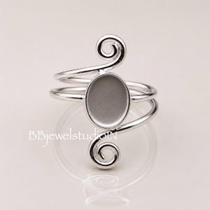 Peut inclure: Une bague argentée avec un sertissage ovale et un design tourbillonnant. La bague présente un double anneau et des spirales décoratives en haut et en bas. Le texte "BBjewelstudioN" est visible en bas à gauche.