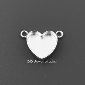 May include: A silver heart-shaped jewellery finding with two small loops on either side. The centre of the heart is a recessed area. The text "BB Jewel Studio" is printed below the heart.