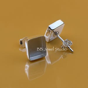 Könnte beinhalten: Zwei quadratische Ohrstecker aus Silber mit Stecker und Butterfly-Verschluss. Die Ohrringe haben ein ausgehöhltes quadratisches Design, ideal zum Setzen von Steinen oder anderen Verzierungen. Die Ohrringe werden auf einer reflektierenden, goldbraunen Oberfläche präsentiert. Der Text "BB Jewel Studio" ist sichtbar.